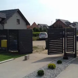 Nowoczesna, pozioma brama wjazdowa z brązowych paneli, częściowo otwarta, z widokiem na podjazd i biały samochód w tle, na tle domów jednorodzinnych.