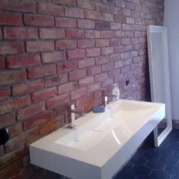 Nowoczesna, prostokątna umywalka z białego konglomeratu zawieszona na ceglanej ścianie, z widoczną baterią, mydłem i fragmentem czarnej, heksagonalnej podłogi.