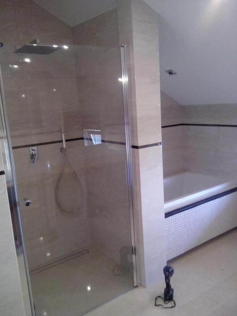 Nowoczesna łazienka z kabiną prysznicową typu walk-in, wykończona beżowymi płytkami z czarnym paskiem dekoracyjnym i białą wanną obłożoną mozaiką. Na podłodze leży narzędzie.