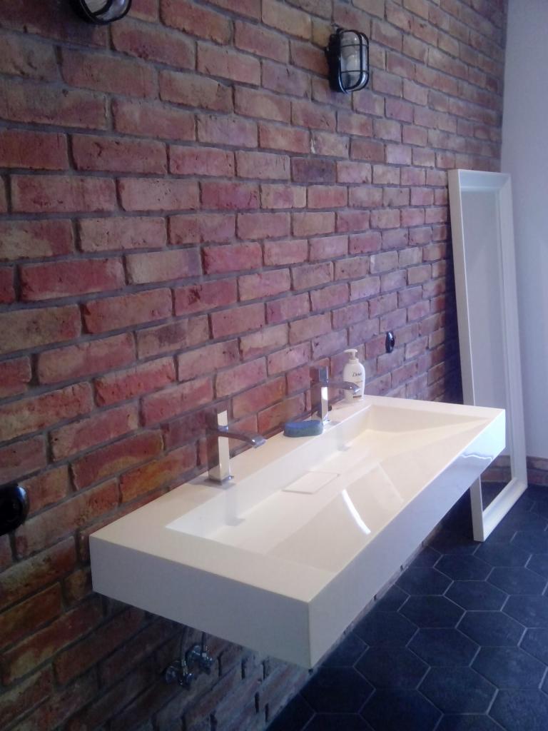 Nowoczesna, prostokątna umywalka z białego konglomeratu zawieszona na ceglanej ścianie, z widoczną baterią, mydłem i fragmentem czarnej, heksagonalnej podłogi.