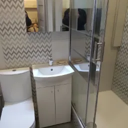 Nowoczesna łazienka z białą umywalką z szafką, toaletą i kabiną prysznicową ze szklanymi drzwiami, wykończona płytkami w zygzaki w odcieniach szarości i bieli.