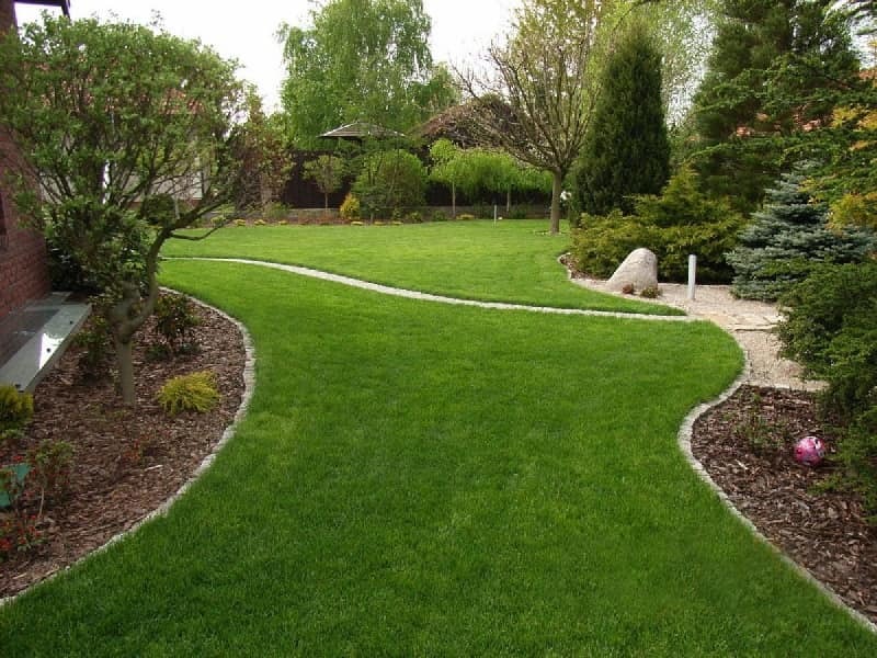 Zadbany trawnik z krętą ścieżką obramowaną kamieniami, prowadzącą przez zielony ogród z drzewami i krzewami. W tle widoczna altana i dom.