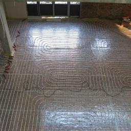 Dariusz Szaferski - Instalacje, Wykończenia i Remonty - Widok z góry na rozłożone rury systemu ogrzewania podłogowego na siatce montażowej, częściowo zalane wylewką, z widocznym słupem konstrukcyjnym i oknami w tle.