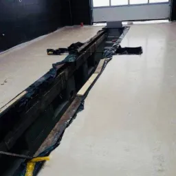 Świeżo wylana, jasnoszara posadzka w hali przemysłowej z widocznym kanałem rewizyjnym zabezpieczonym folią i deskami. W tle brama garażowa i czarne ściany.