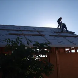 Prace dekarskie na dachu w trakcie budowy. Widoczni dwaj dekarze montujący izolację na drewnianej konstrukcji. Ujęcie z dołu, widok na błękitne niebo.