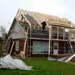 Dom w trakcie budowy dachu, widoczne rusztowania, drewniane elementy konstrukcyjne i pracownik na górze. Na pierwszym planie trawa i materiały budowlane.