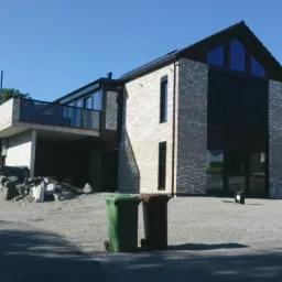 Nowoczesny dom jednorodzinny z ceglaną elewacją, dużymi oknami i balkonem, widoczny z poziomu ulicy z dwoma pojemnikami na śmieci na pierwszym planie.