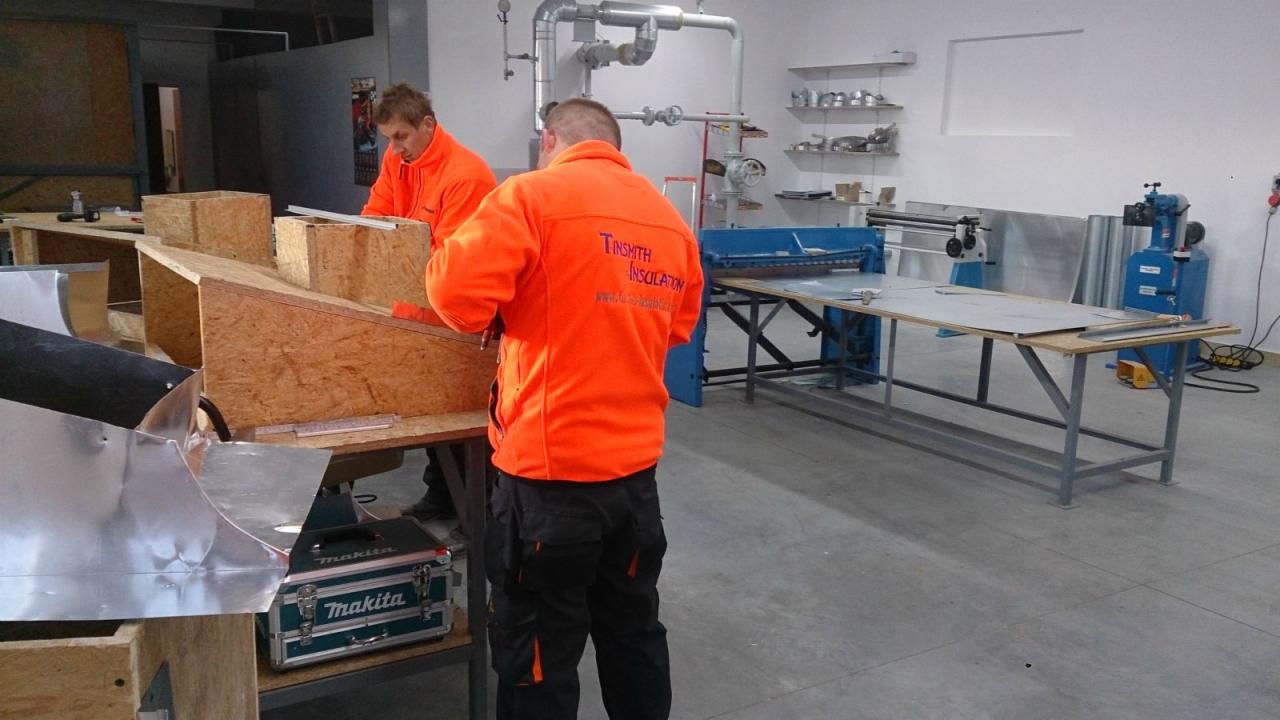 Dwóch monterów w pomarańczowych uniformach pracuje nad konstrukcją z blachy w warsztacie, używając narzędzi; widoczna skrzynka narzędziowa Makita i maszyny blacharskie.