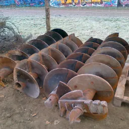 Stos metalowych świdrów do gruntu ułożonych na ziemi, obok żółty element maszyny budowlanej, w tle trawa pokryta szronem i graffiti na ogrodzeniu.