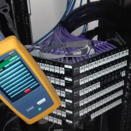 Tester okablowania Fluke Networks podłączony do panelu krosowniczego z fioletowymi kablami, obok leży czołówka Petzl.