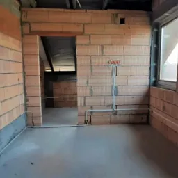 Wnętrze domu w stanie surowym: ściany z czerwonej cegły dziurawki, betonowa wylewka, okna PCV z widokiem na zabudowę jednorodzinną, instalacja elektryczna w peszlach na ścianie.