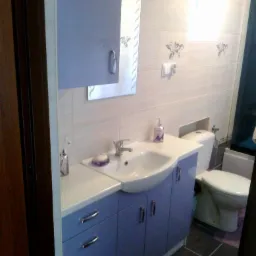 Niebieska szafka łazienkowa z umywalką i lustrem w komplecie, widok na toaletę i fragment drzwi. Ukończona aranżacja łazienki, płytki na ścianach i podłodze.