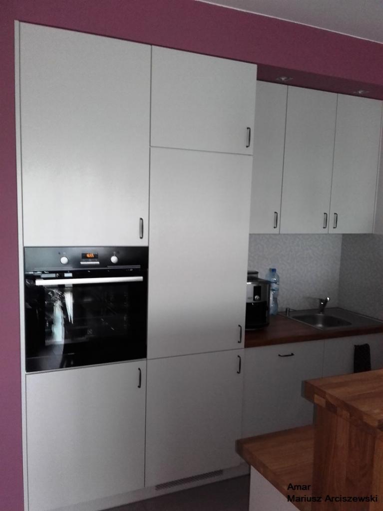 Jasnoszara zabudowa kuchenna z piekarnikiem w ścianie, minimalistyczny design, fioletowa ściana w tle. Widoczny fragment blatu i zlewu.