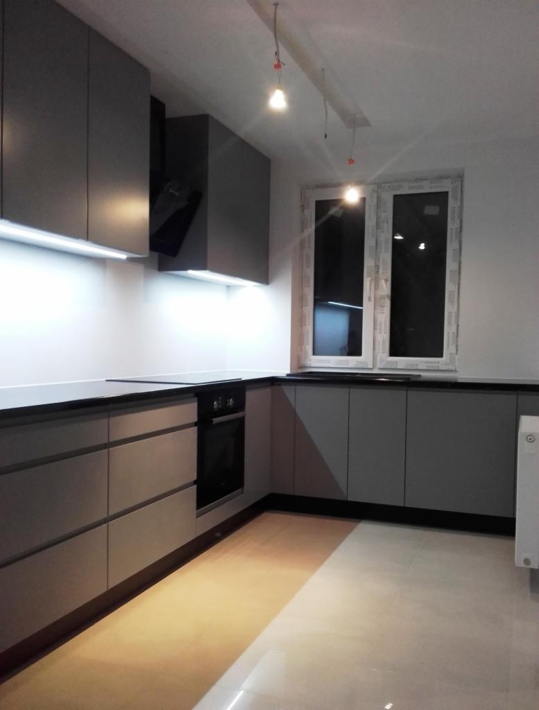 Szara, minimalistyczna kuchnia z czarnym blatem i oświetleniem LED pod szafkami. Widoczny piekarnik i okap. Jasne płytki na podłodze.