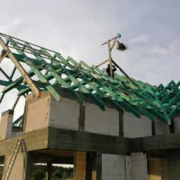 Surowy stan domu z betonu komórkowego z częściowo ukończoną, zieloną konstrukcją drewnianą dachu i wieńcem z kwiatów zawieszonym na szczycie, drabina przystawiona do ściany.