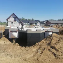 Fundament domu w trakcie budowy, zabezpieczony czarną izolacją, z widocznymi białymi elementami szalunkowymi, na tle wykopu i częściowo wybudowanego domu z białego pustaka.