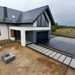 Nowoczesny dom jednorodzinny z ciemnoszarą dachówką, antracytowymi oknami i bramą garażową, jasną elewacją. Ułożona kostka brukowa w odcieniach szarości, widoczne materiały budowlane i krajobraz...