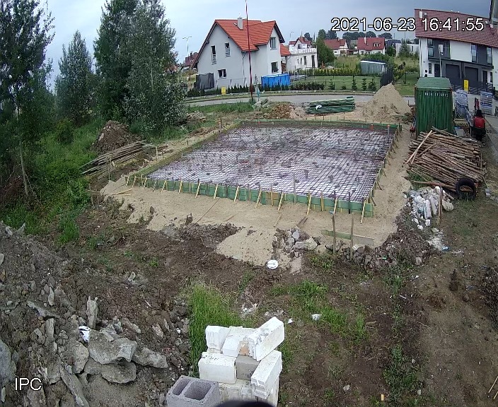 Widok z góry na przygotowany teren pod fundamenty z ułożonym zbrojeniem i szalunkiem, widoczna data i godzina na zdjęciu, w tle zabudowa jednorodzinna.