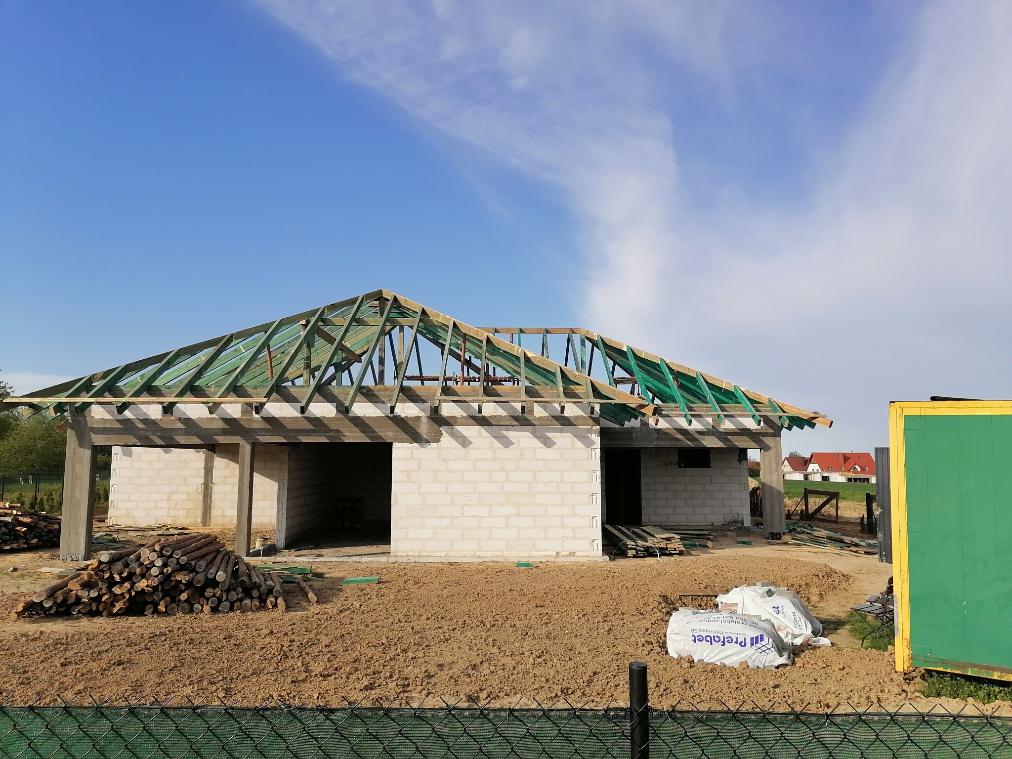 Budowa domu jednorodzinnego w stanie surowym otwartym, widoczne ściany z bloczków, drewniana konstrukcja dachu z zieloną membraną, stos drewna na pierwszym planie, w tle inne domy.