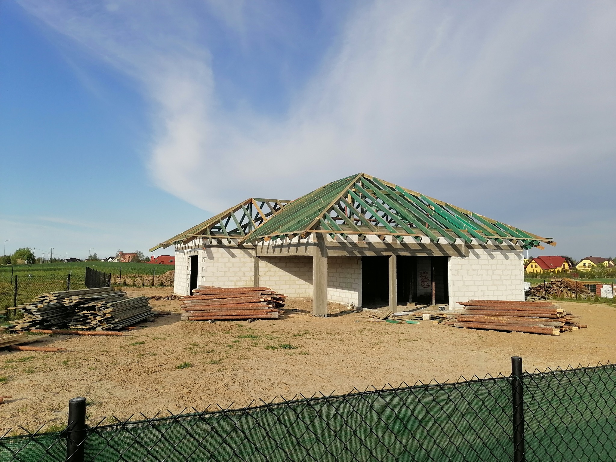 Niedokończony dom z białego pustaka z widoczną konstrukcją dachu z zielonych łat, stosy drewna na placu budowy, widok zza zielonego ogrodzenia siatkowego.