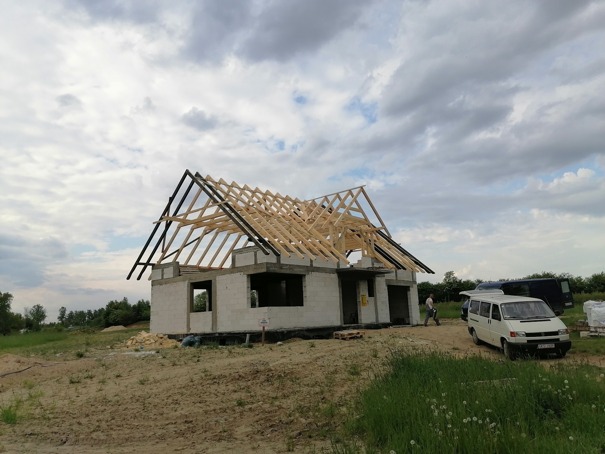 Niedokończony budynek z pustaków z drewnianą konstrukcją dachu, na zewnątrz zaparkowany biały van, pochmurne niebo w tle.