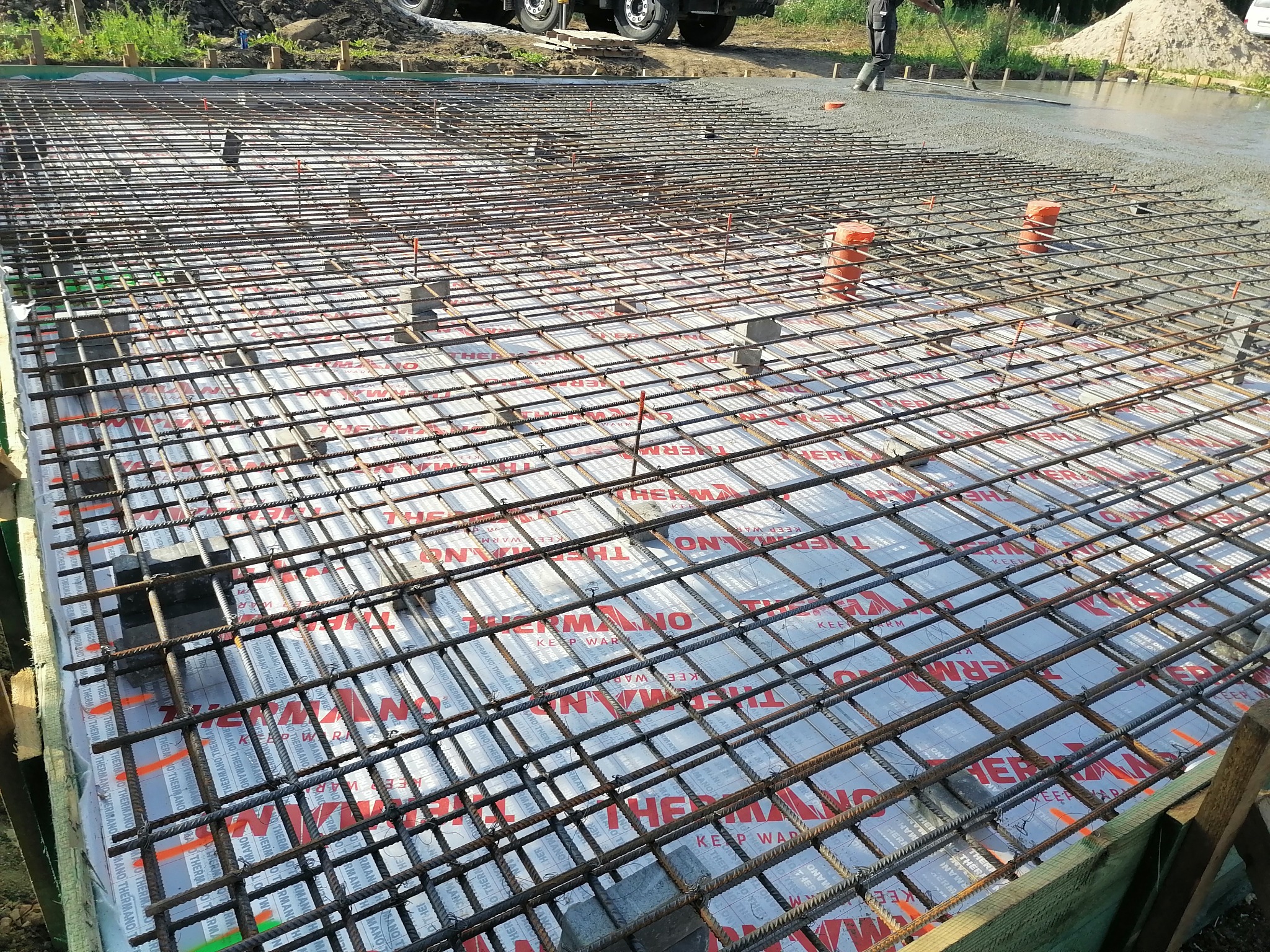 Zbrojenie płyty fundamentowej z siatki stalowej na izolacji termicznej, przygotowane do zalania betonem, z widocznym pracownikiem wyrównującym powierzchnię betonu w tle.