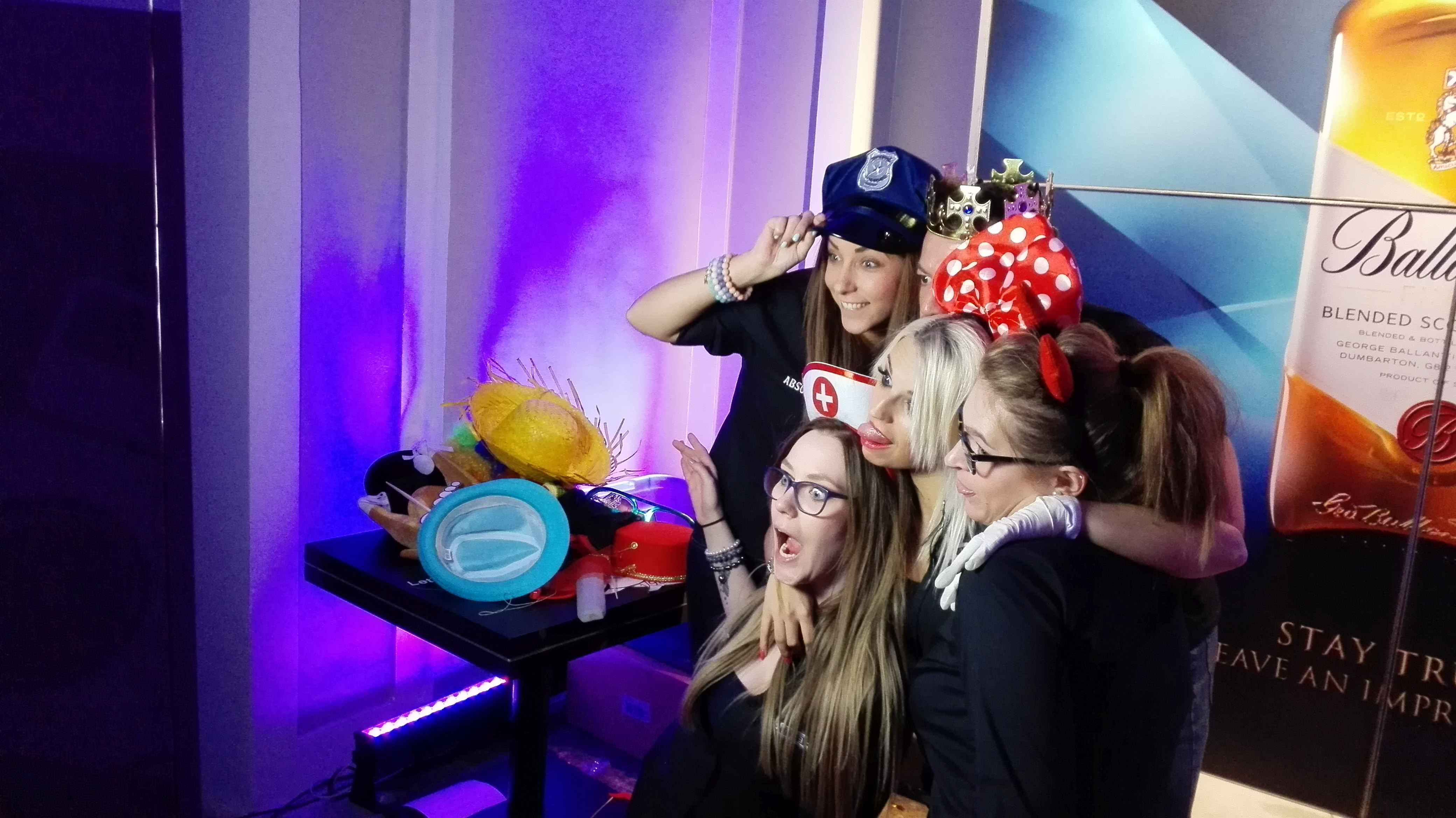 Grupa kobiet w przebraniach pozuje do zdjęcia na tle fotobudki z rekwizytami w Warszawie. Uśmiechnięte twarze i zabawne nakrycia głowy.