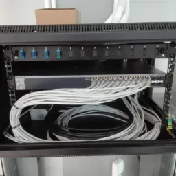 Szafa rack z zamontowanym switchem Netgear i okablowaniem sieciowym, widoczny panel krosowniczy z niebieskimi przełącznikami oraz uziemienie.