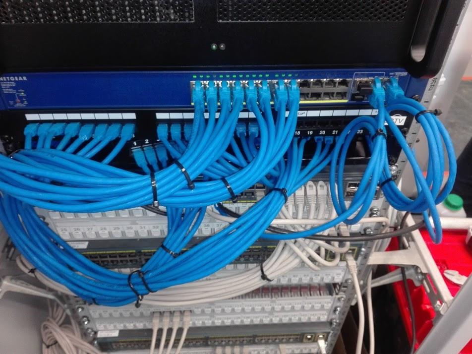 Rządowe szafy rack z niebieskimi i białymi kablami sieciowymi uporządkowanymi za pomocą czarnych opasek zaciskowych, widoczne porty i oznaczenia numerów.