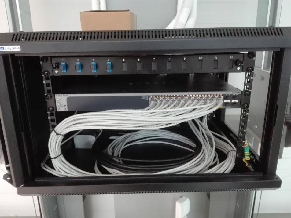 Szafa rack z zamontowanym switchem Netgear i okablowaniem sieciowym, widoczny panel krosowniczy z niebieskimi przełącznikami oraz uziemienie.