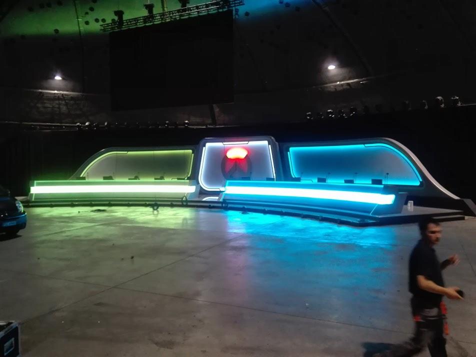 Nowoczesna, podświetlana konstrukcja sceniczna w przestronnym, ciemnym pomieszczeniu, z widoczną osobą i fragmentem samochodu w tle.