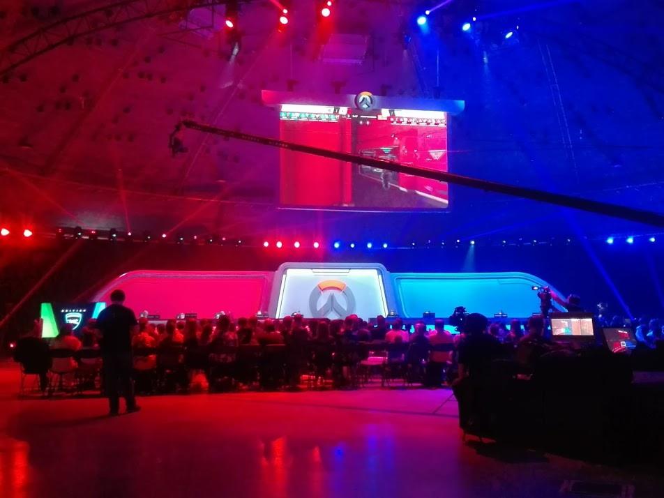 Szeroki plan hali widowiskowej oświetlonej czerwonym i niebieskim światłem, z widoczną sceną podczas turnieju e-sportowego, na której widać telebim z grą, a przed sceną siedzą widzowie.