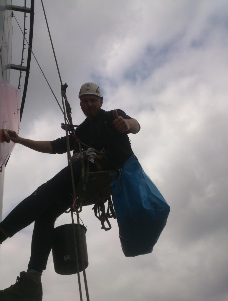 Pracownik w uprzęży, z kaskiem na głowie, daje kciuk w górę podczas czyszczenia elewacji budynku na wysokości, z widocznym wiadrem i niebieską torbą na narzędzia.