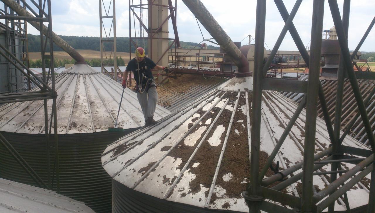 Pracownik w uprzęży, z żółtym kaskiem, czyści metalową powierzchnię dachu silosu za pomocą szczotki na długim kiju, widoczne zanieczyszczenia na dachu, konstrukcja wsporcza i krajobraz w tle.