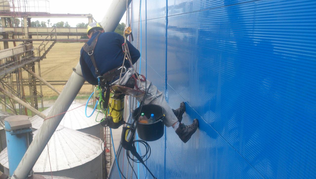 Pracownik w uprzęży, z wiadrem i myjką ciśnieniową, czyści niebieską, pionową ścianę silosu zbożowego na wysokości.