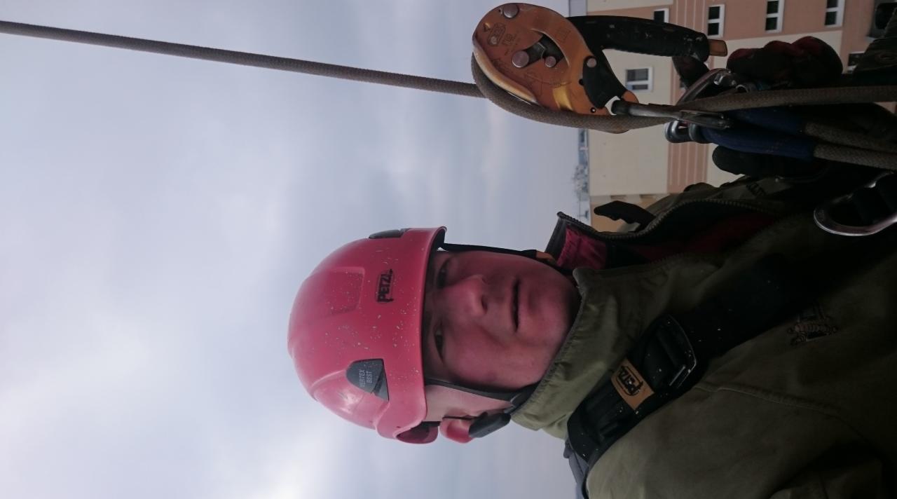 Pracownik w czerwonym kasku Petzl, zabezpieczony linami, w trakcie pracy na wysokości z widokiem na budynek mieszkalny i pochmurne niebo.