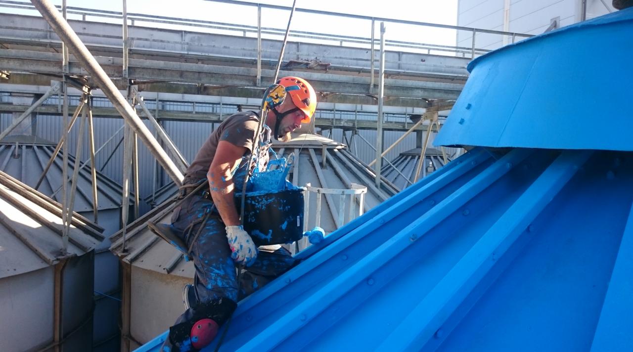 Pracownik w kasku i uprzęży maluje na niebiesko dach silosu zbożowego, widoczne ślady farby na ubraniu i rękawicach, metalowa konstrukcja w tle.