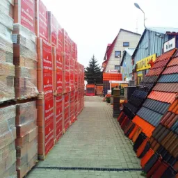 Skład budowlany: palety z cegłami Porotherm i ekspozycja dachówek w różnych kolorach. Widok wzdłuż alejki, na zewnątrz, w dzień.