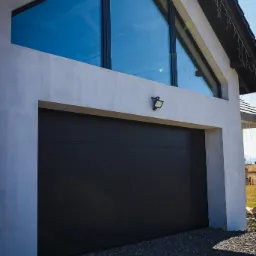 Nowoczesny dom z szarą elewacją, dużą bramą garażową w kolorze antracytowym i trójkątnym oknem na poddaszu, oświetlony lampą zewnętrzną. Widoczny fragment dachu z dekoracyjnym oświetleniem.