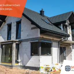 Dom w trakcie budowy z widocznymi oknami, roletami zewnętrznymi i izolacją fundamentów, białe bloczki konstrukcyjne, ciemna dachówka, błękitne niebo, częściowo ocieplony budynek, materiały...