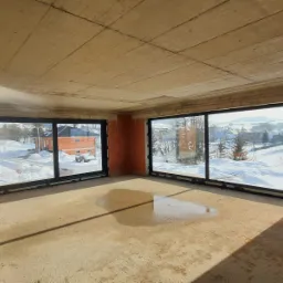 Surowy stan wnętrza budynku w trakcie budowy z zamontowanymi dużymi oknami z czarnymi ramami, widok na zaśnieżony krajobraz za oknem, na podłodze kałuża wody.