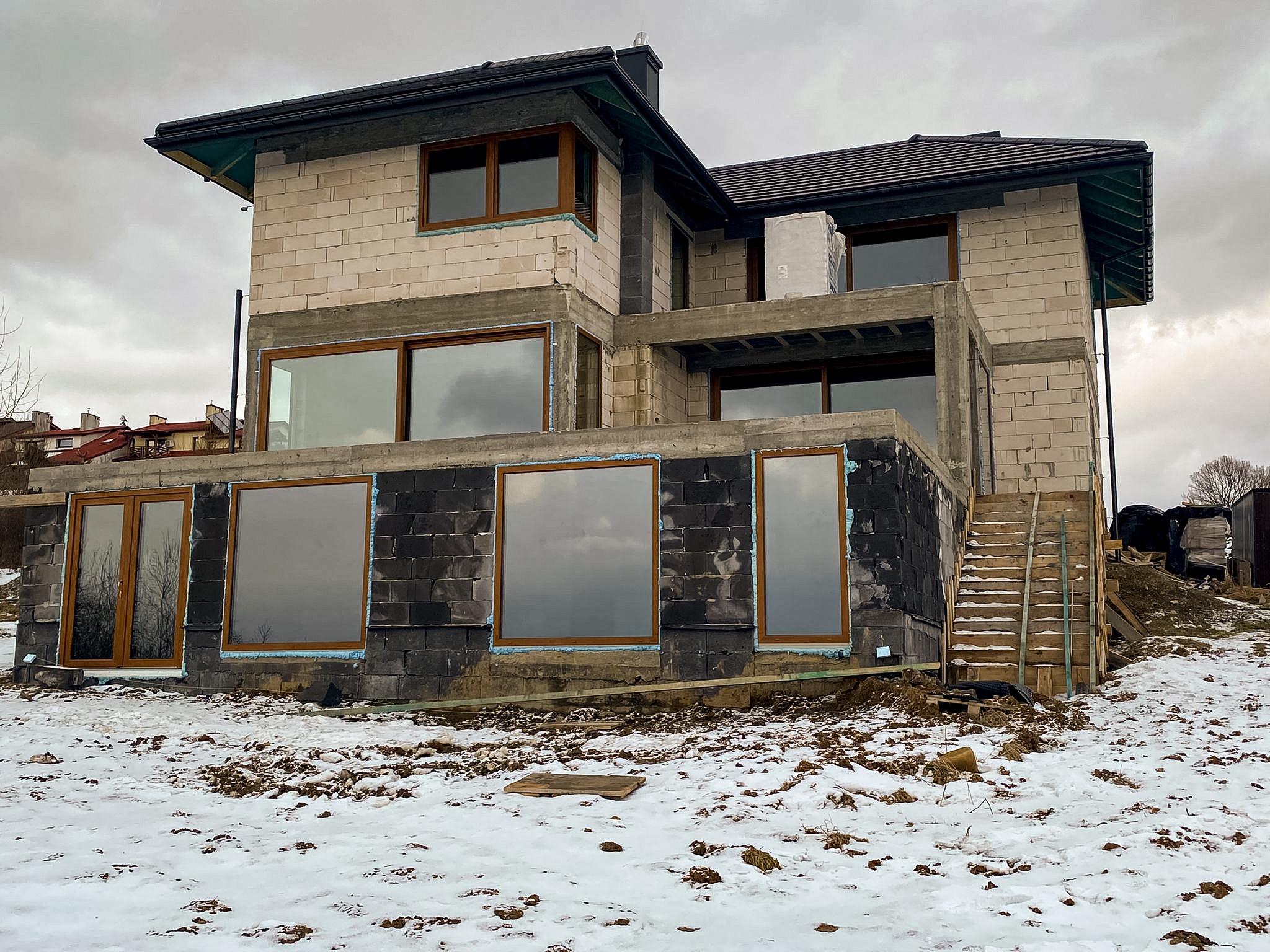 Surowy stan dwukondygnacyjnego domu z oknami z brązowymi ramami, częściowo ocieplonymi fundamentami, widoczne drewniane schody i śnieg na ziemi.