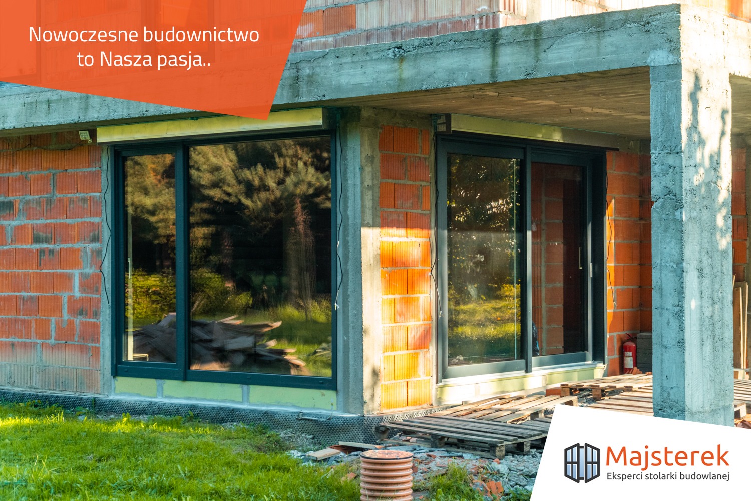 Nowe okna przesuwne z ciemną ramą w surowym stanie budynku, widoczne cegły i betonowe filary, teren budowy z paletami i trawą.