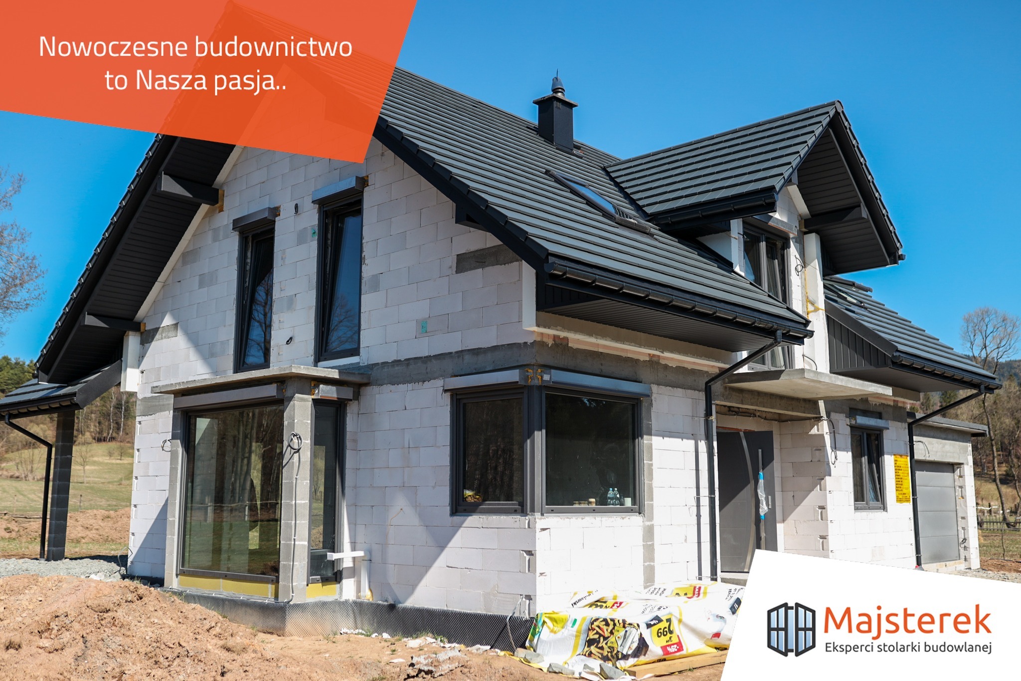 Dom w trakcie budowy z widocznymi oknami, roletami zewnętrznymi i izolacją fundamentów, białe bloczki konstrukcyjne, ciemna dachówka, błękitne niebo, częściowo ocieplony budynek, materiały...