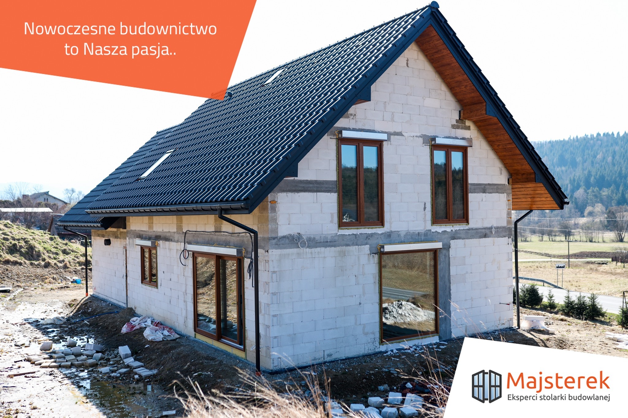 Budowa domu jednorodzinnego w Żywcu w stanie surowym otwartym, widoczne ściany z bloczków, zamontowane okna z brązowymi ramami, czarny dach z blachy, teren wokół domu częściowo uporządkowany.