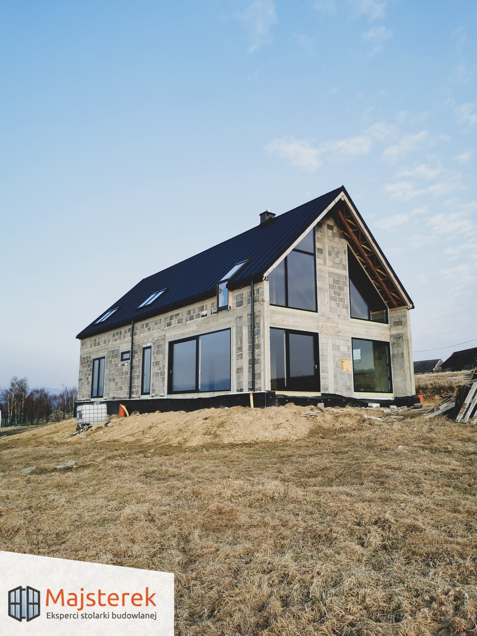 Surowy dom z szarego betonu komórkowego z zamontowanymi dużymi, czarnymi oknami aluminiowymi, na etapie budowy, widoczne elementy izolacji fundamentów i czarne pokrycie dachu.