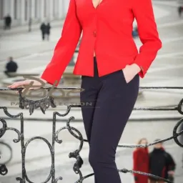 Kobieta w czerwonej marynarce i granatowych spodniach pozuje na tle miejskiego placu, opierając się o ozdobną metalową barierkę.