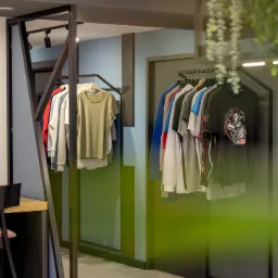 Wnętrze butiku odzieżowego z ubraniami na wieszakach, widoczne koszulki z nadrukami i jednolite, minimalistyczny design, ciemne metalowe ramy ekspozycyjne, oświetlenie punktowe, dekoracyjne rośliny...