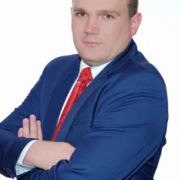 Portret mężczyzny w niebieskim garniturze i czerwonym krawacie, z założonymi rękami na białym tle.