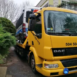 Ż&oacute;łta laweta DAF z numerem telefonu 504 504 112 przewozi policyjny samoch&oacute;d dostawczy na tle budynku mieszkalnego z balkonem i zielenią.